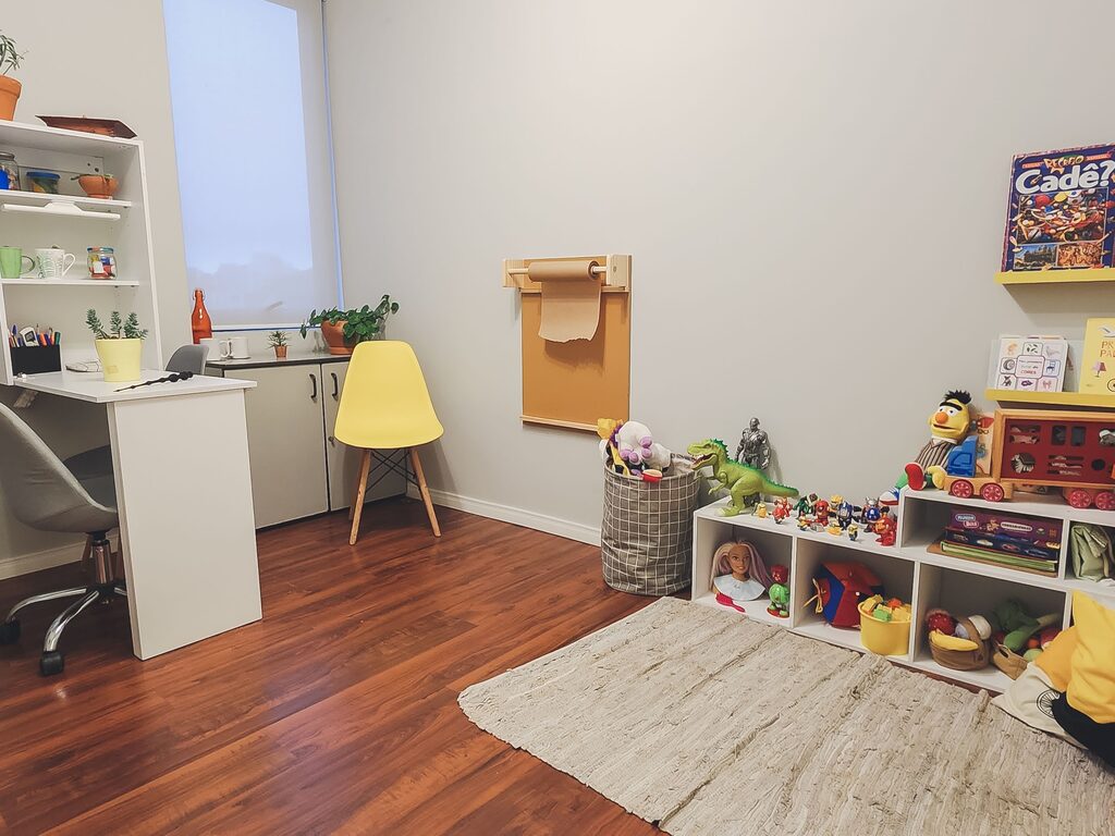 Sala infantil da clínica, com mesa de atividades, cadeira amarela, tapete e estante de brinquedos.